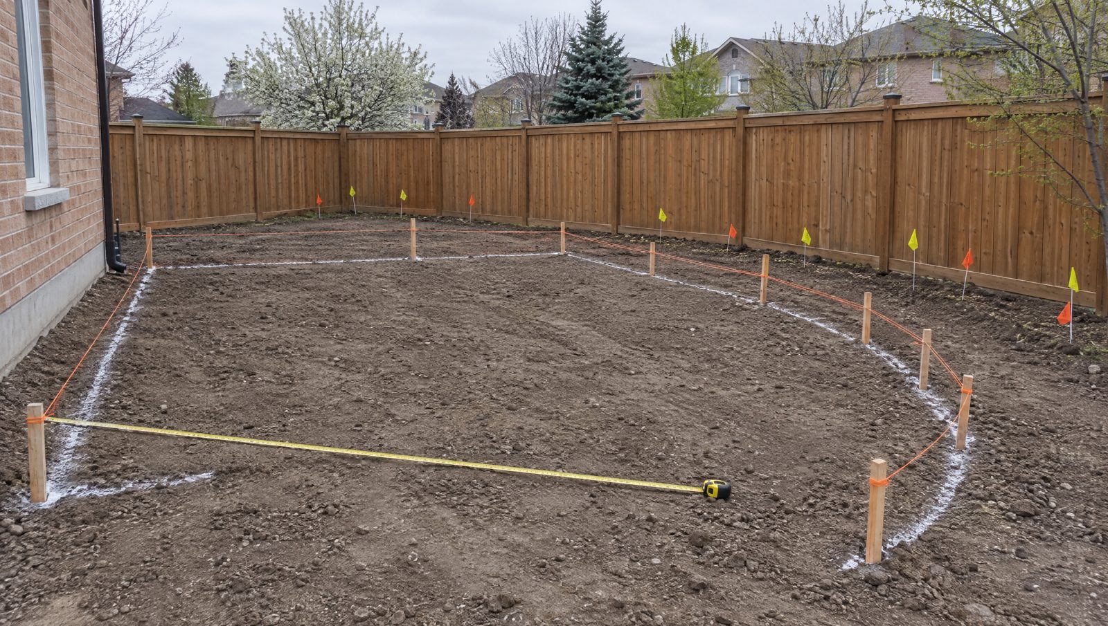 A bare backyard staked out with orange string lines, white paint marks and small utility locate flags