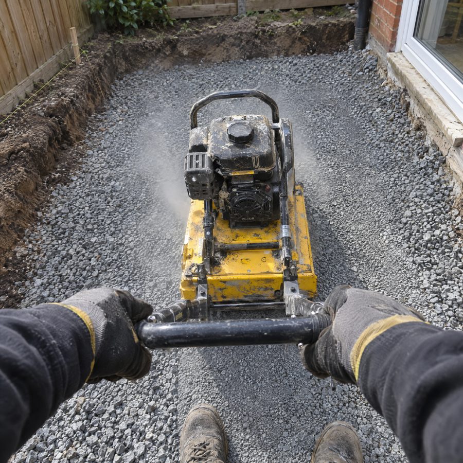Gloved hands guiding a plate compactor across a gravel base, string line and wooden stakes in the foreground